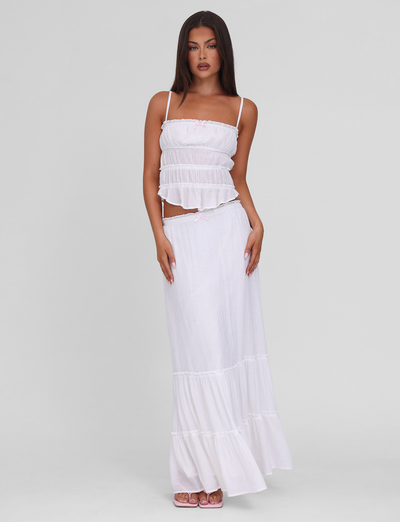 A woman with long brown hair wears the TIGERMIST ASHLYNN MAXI SKIRT in white—a tiered, elastic-waist maxi skirt—paired with a white sleeveless ruffled crop top. She stands against a light gray background, looking forward with a neutral expression.