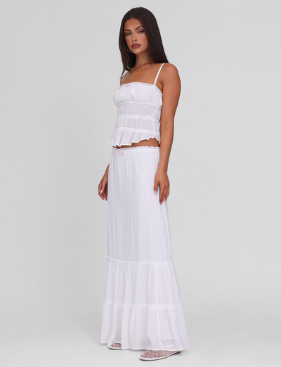 A woman with long dark hair wears a sleeveless white crop top and the TIGERMIST ASHLYNN MAXI SKIRT in white, featuring an elasticated waist. She stands against a plain light gray background, facing slightly to the side.