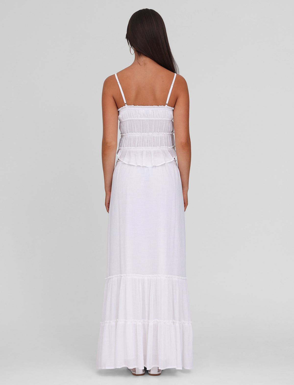 A woman with long brown hair, seen from behind, wears the TIGERMIST ASHLYNN MAXI SKIRT in white, featuring an elasticated waist and ruffle detailing. She stands against a plain light gray background.
