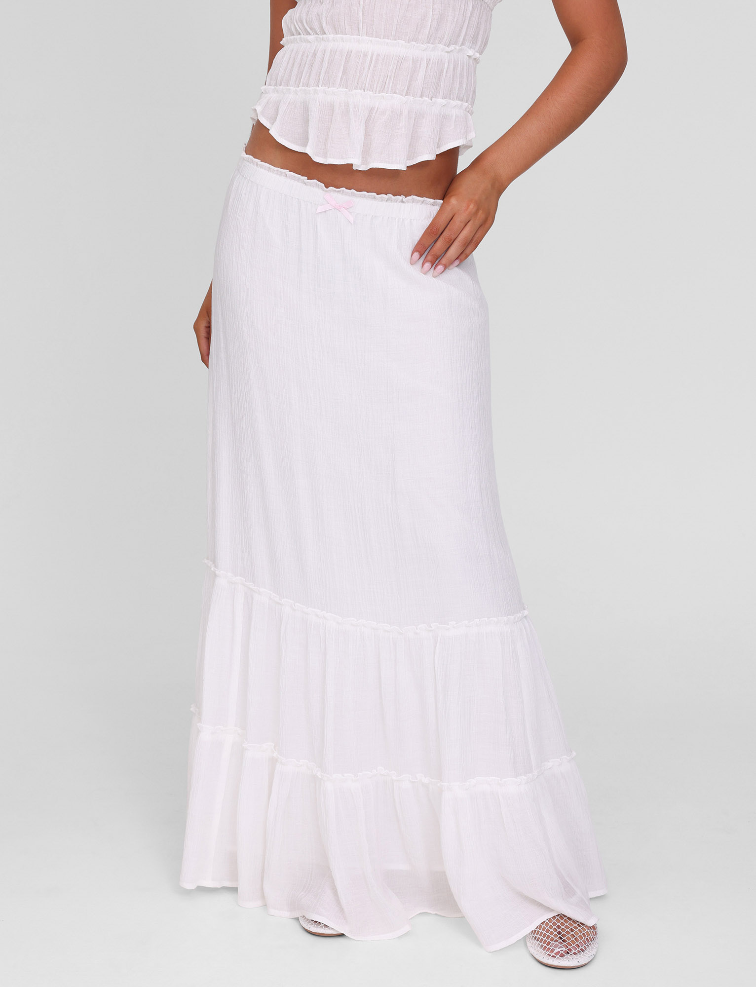 A woman models the TIGERMIST ASHLYNN MAXI SKIRT in white, featuring a tiered design and elasticated waist, paired with a matching sleeveless top; her head is out of frame and she rests a hand on her hip against a plain light background.