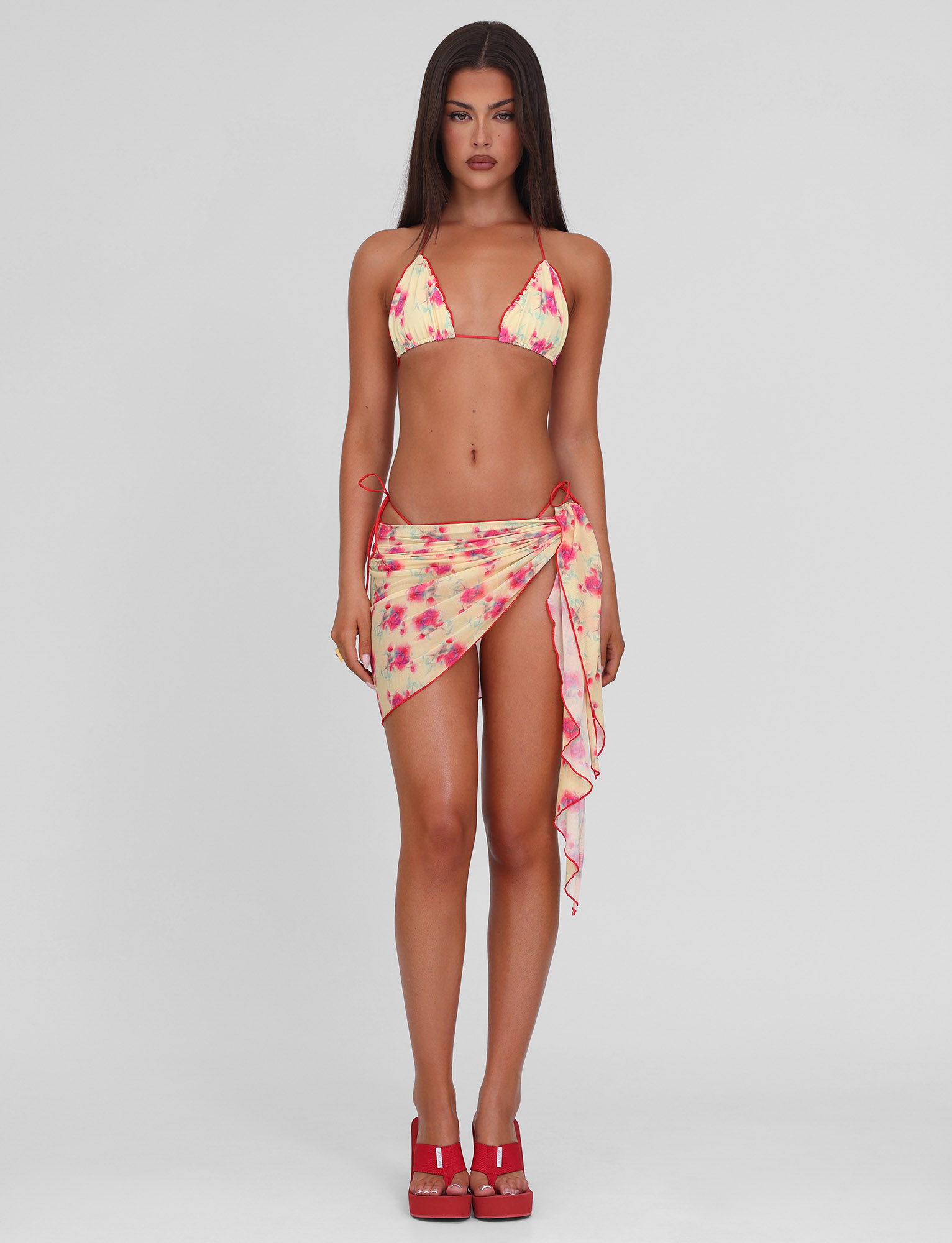 A woman with long brown hair models the TIGERMIST AMALIE 2PC BIKINI BOTTOM - SUMMER BLOSSOM set and matching sarong, paired with red platform heels, against a plain white background.