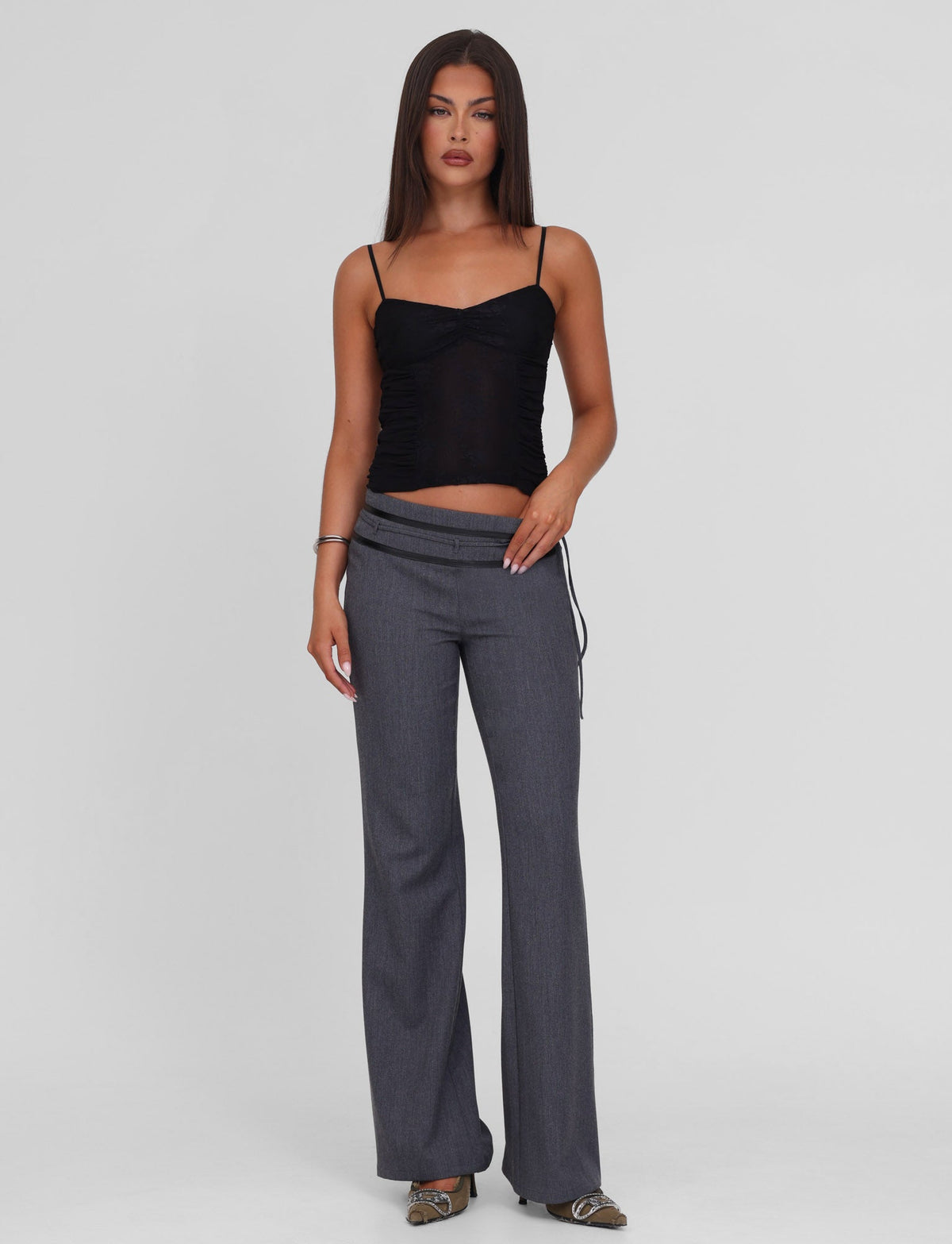 A woman with straight brown hair and neutral makeup wears the TIGERMIST KALLIE TOP - BLACK, a lace camisole with adjustable straps, paired with gray low-rise wide-leg trousers featuring a loose belt detail against a plain background.