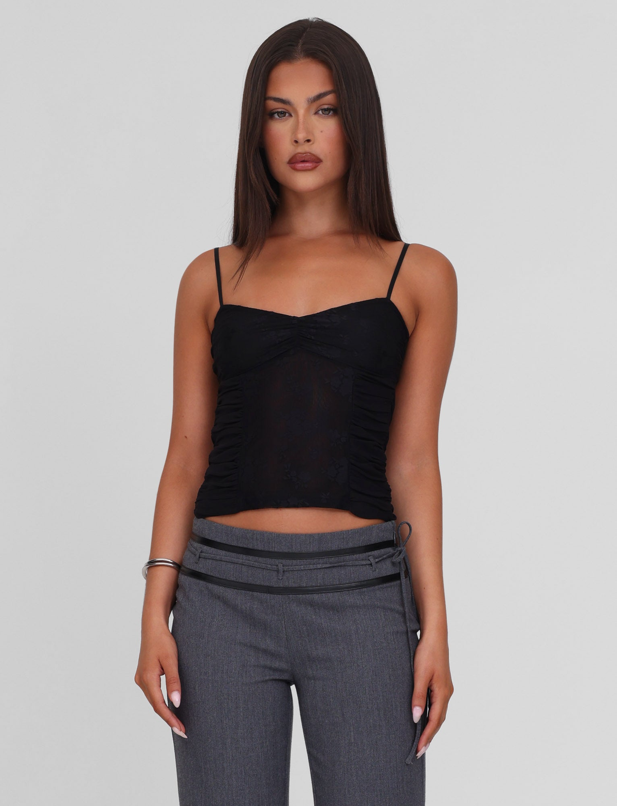 A woman with straight dark hair models the TIGERMIST KALLIE TOP - BLACK, a black lace camisole with adjustable straps, paired with gray pants against a plain light background, looking directly at the camera with a neutral expression.