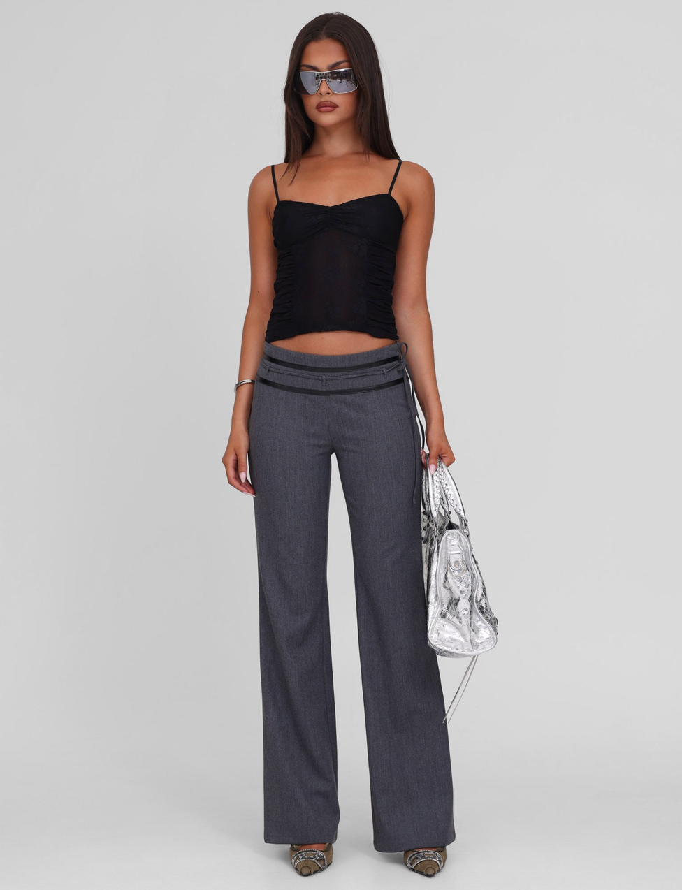 A woman wearing the TIGERMIST KALLIE TOP - BLACK, gray wide-leg trousers with an asymmetric waistband, metallic heels, sunglasses, and carrying a shiny silver handbag poses against a plain light gray background.