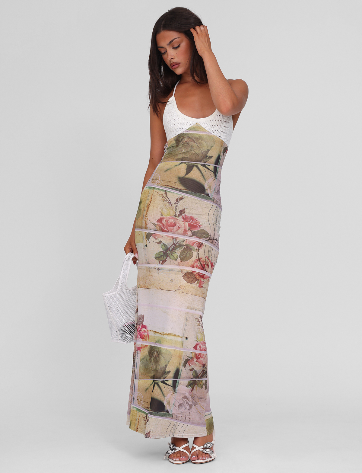 A woman poses in the TIGERMIST TILLEY MAXI DRESS - ROSE POSTCARD, a sleeveless white maxi dress with an exclusive floral print. She holds a white mesh handbag, wears white sandals, and stands against a plain light gray background.