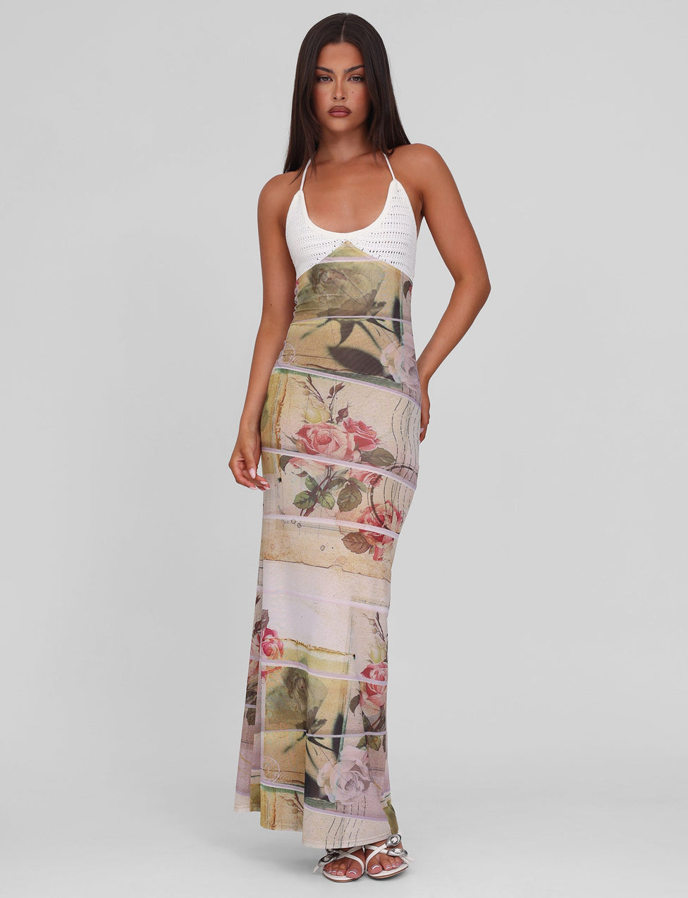 A woman with long, straight hair wears the TIGERMIST TILLEY MAXI DRESS - ROSE POSTCARD, featuring a fitted printed mesh, contrast crochet bust cups, and pastel floral skirt. She stands on a plain background in silver sandals.