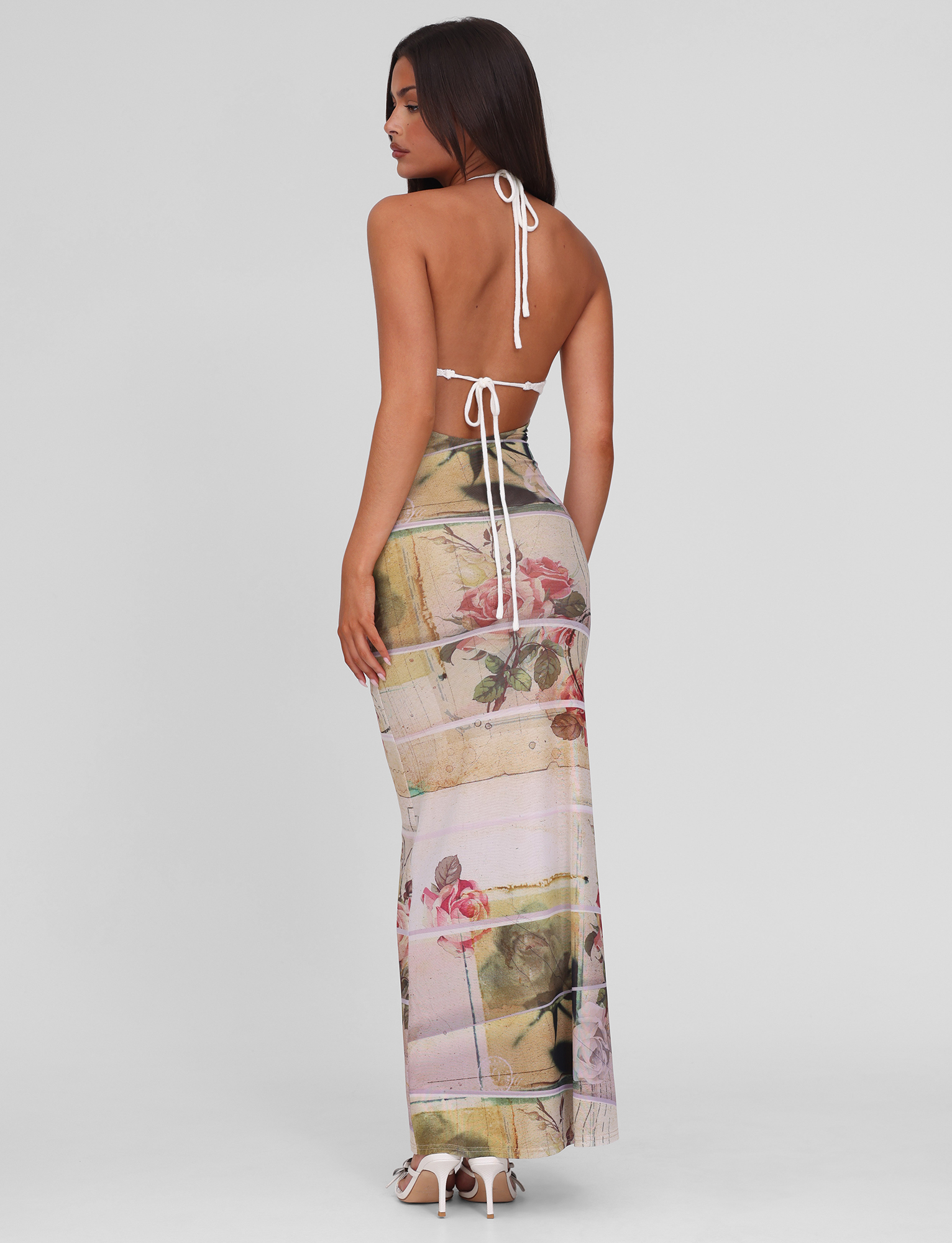 A woman faces away in the TIGERMIST TILLEY MAXI DRESS - ROSE POSTCARD, featuring an exclusive print and crochet bust cups. She pairs it with white heeled sandals and has long straight hair, set against a plain light background.