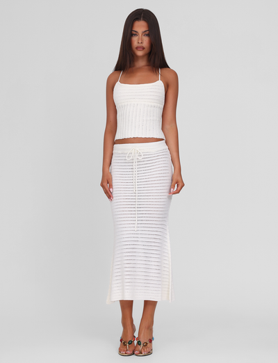 A woman stands against a plain white background wearing the TIGERMIST EMELYN TOP in cream, a sleeveless crochet crop with lace-up open back, paired with a matching ankle-length skirt and sandals.
