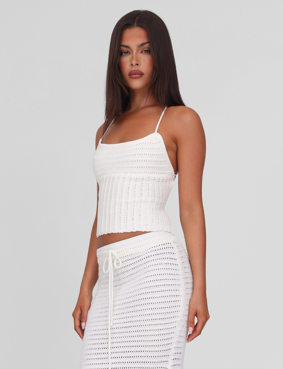 A woman with long dark hair wears the EMELYN TOP in cream by TIGERMIST, featuring thin straps and an open back. She stands against a plain light background, looking confidently at the camera.