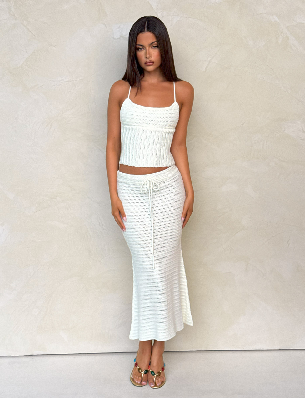 A woman with long dark hair and open-toed heeled sandals stands against a textured beige wall, wearing a sleeveless white knit top and the TIGERMIST EMELYN MIDI SKIRT in cream with an elasticated waist.