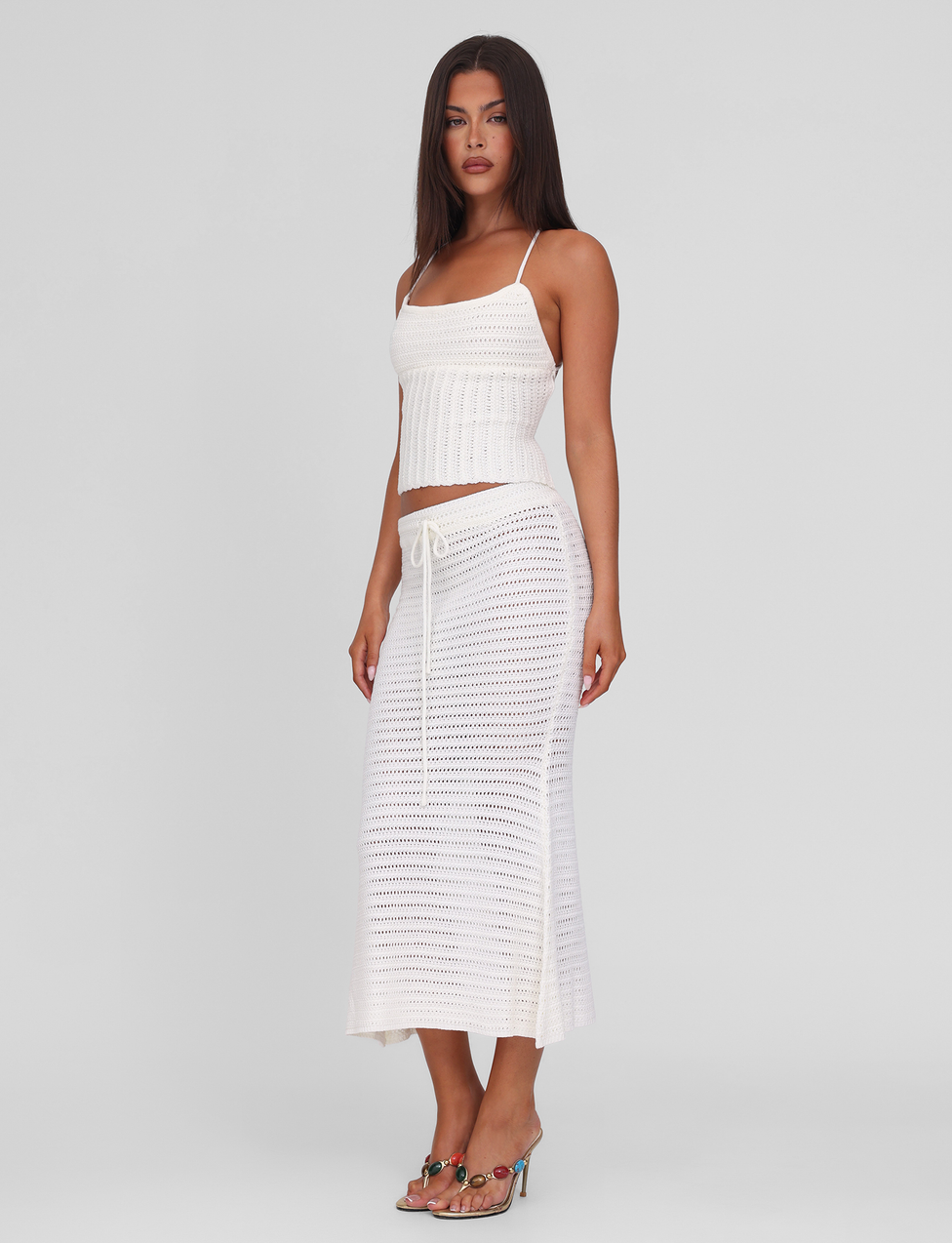 A woman in heeled sandals wears the EMELYN MIDI SKIRT - CREAM by TIGERMIST, a semi-sheer crochet midi skirt with an elasticated waist, paired with a white crochet tank top against a plain light background.