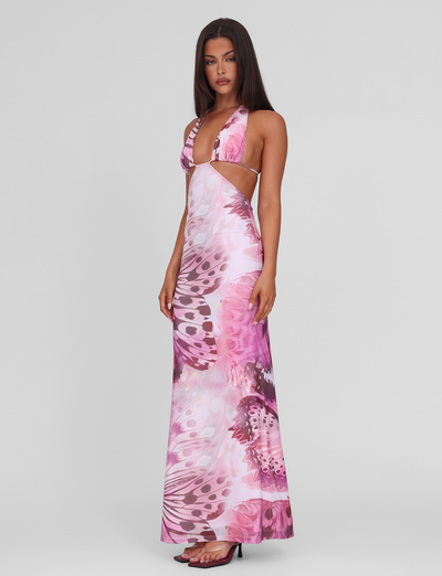 A woman wears the TIGERMIST SOLEIL MAXI DRESS - MACRO BUTTERFLY, featuring pink and purple butterfly print, adjustable halter ties, side cut-outs, and high stretch fabric. She pairs it with black high-heeled sandals against a white background.