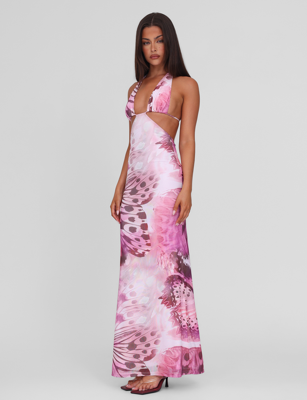 A woman wears the TIGERMIST SOLEIL MAXI DRESS - MACRO BUTTERFLY, featuring pink and purple butterfly print, adjustable halter ties, side cut-outs, and high stretch fabric. She pairs it with black high-heeled sandals against a white background.