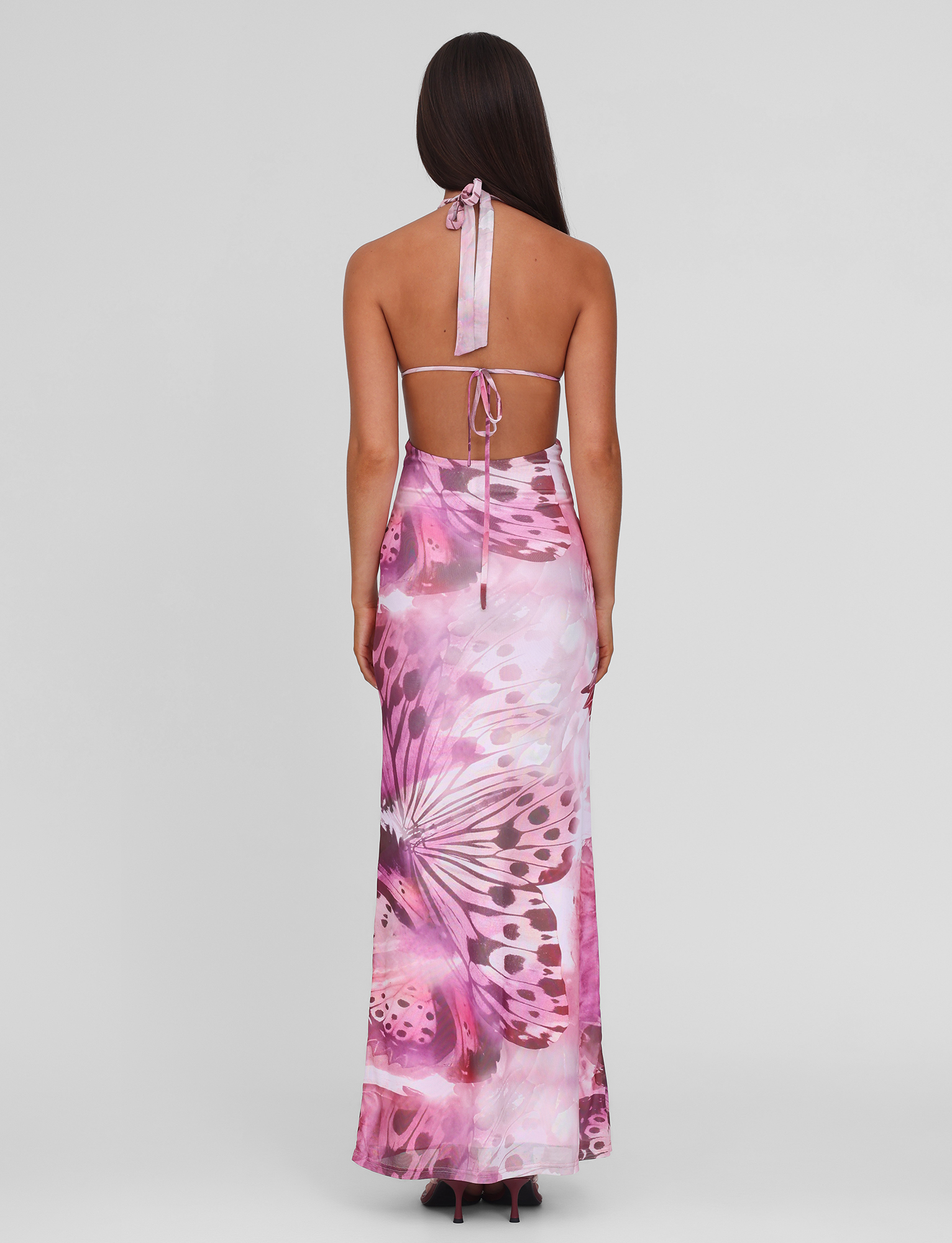 A woman faces away in the TIGERMIST SOLEIL MAXI DRESS - MACRO BUTTERFLY, featuring pink and purple butterfly print, adjustable halter ties, and a backless design. Her long dark hair stands out against the plain white background.