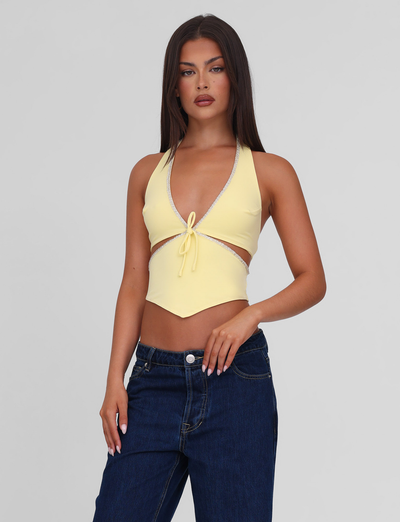A woman with long dark hair wears the TIGERMIST LIZZIE TOP - LEMON, a yellow halter with cut-out details, paired with blue jeans against a plain light gray background.