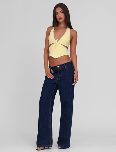 A woman with long dark hair and neutral makeup poses against a plain background in the TIGERMIST LIZZIE TOP - LEMON, a light yellow halter style with beaded trim, paired with dark blue wide-leg jeans. Her right hand rests on her hip.