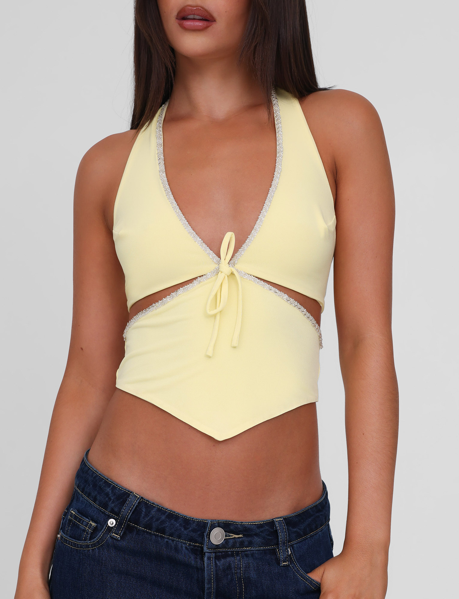 A woman models the TIGERMIST LIZZIE TOP - LEMON, a yellow halter with deep V-neck, beaded trim, and front tie, paired with dark blue jeans. Image is cropped from shoulders to hips.