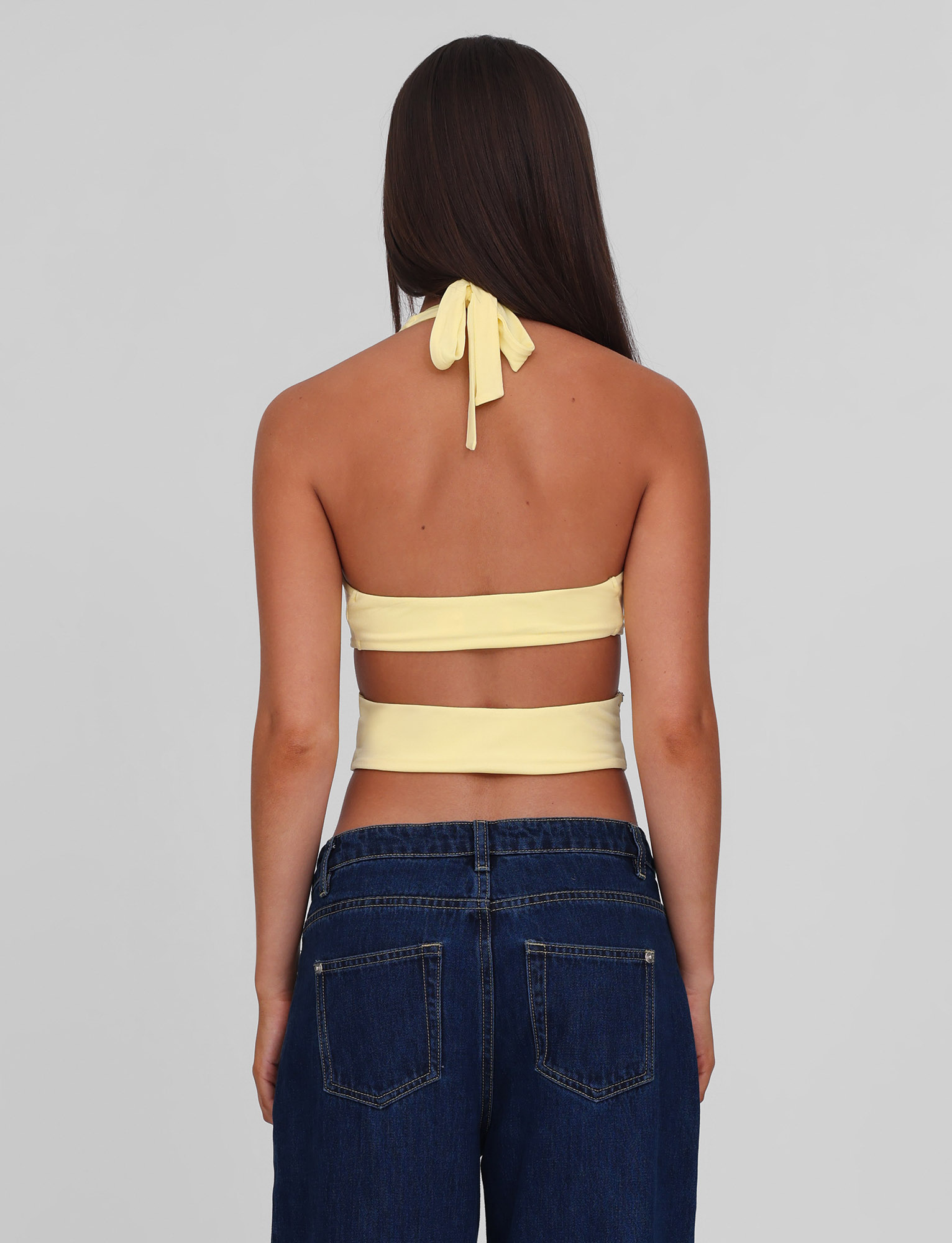 A woman with long brown hair, seen from behind, wears the TIGERMIST LIZZIE TOP - LEMON, a yellow halter top with tie and cut-out details, paired with high-waisted blue jeans against a plain gray background.