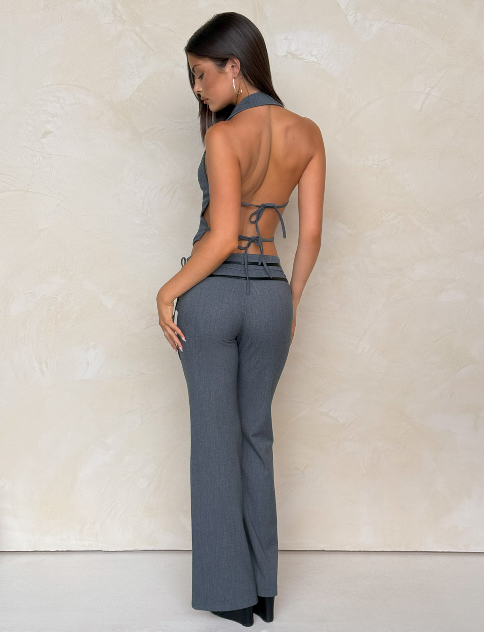 A woman with long dark hair faces away, wearing TIGERMIST's FEDRA PANT in grey—high-waisted suit pants with PU leather trim—and a backless halter top with tie closures, posing against a light textured wall.