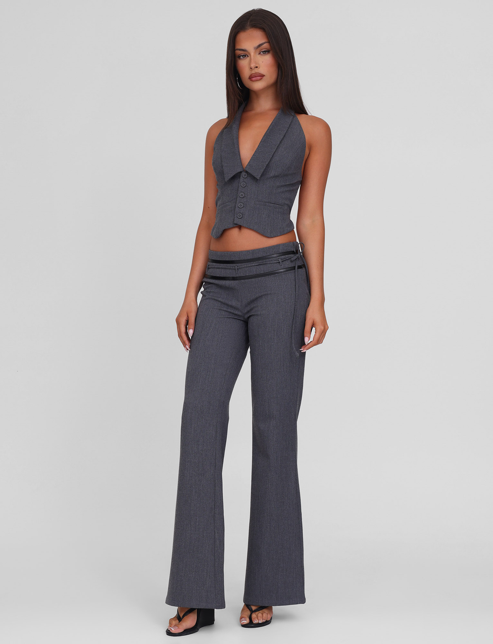 A woman with straight dark hair and a neutral expression wears TIGERMIST's FEDRA PANT - GREY, styled with a sleeveless deep V-neck grey vest featuring PU leather trim and black heeled sandals, against a plain light background.