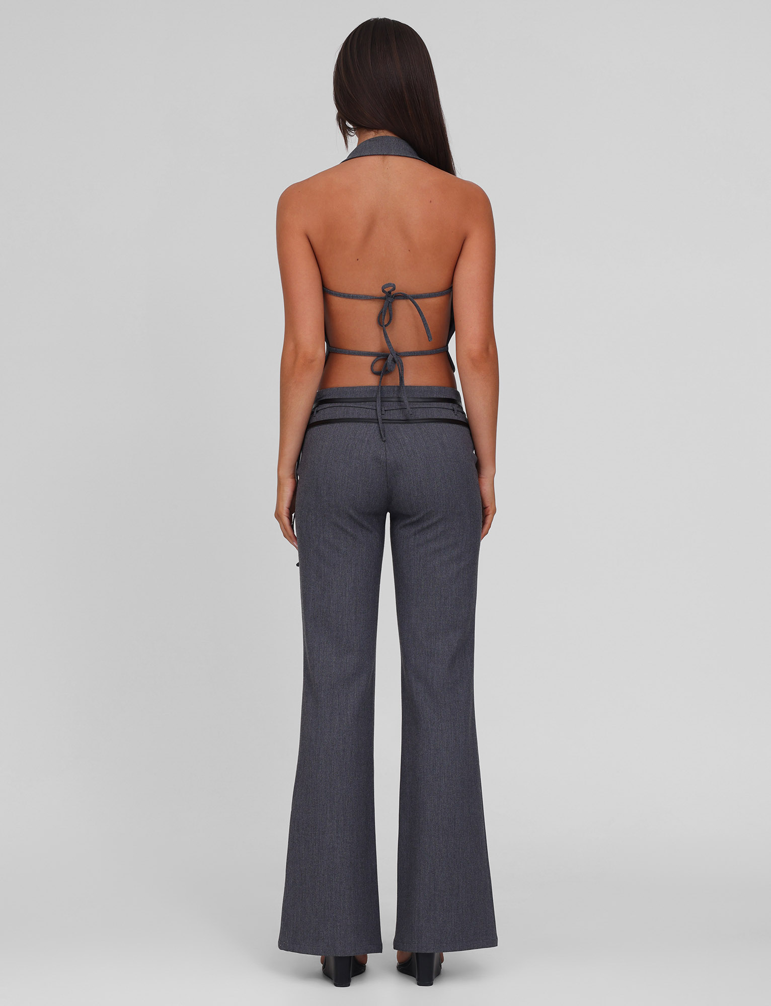 A woman with long straight hair faces away, modeling the TIGERMIST FEDRA PANT in grey and a matching halter-neck top with PU leather trim, tied at the back to reveal her back, against a plain light grey background.