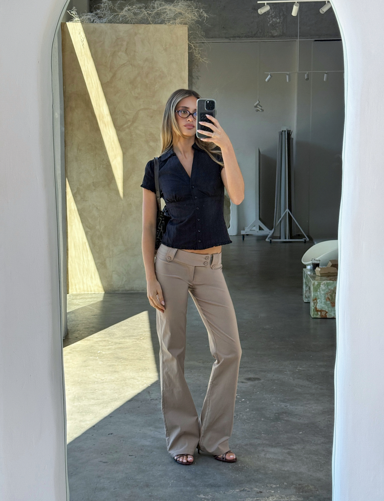 A woman in glasses wears the TIGERMIST VALERIE SHORT SLEEVE SHIRT - BLACK and beige trousers, taking a mirror selfie in a modern, minimalistic room with neutral-toned decor.