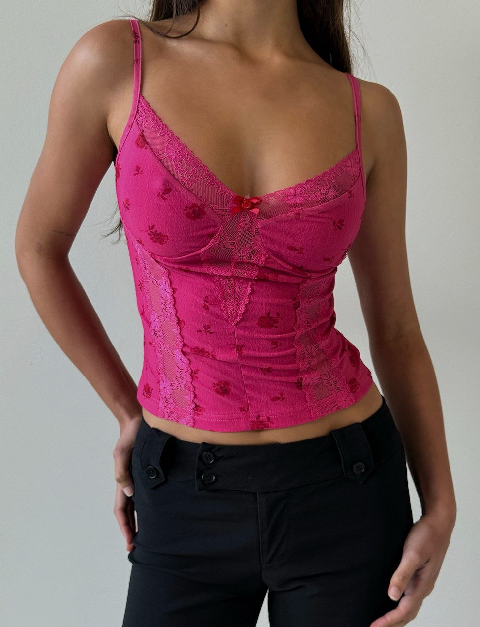A woman wears the TIGERMIST KHALIA TOP - DITSY FLORAL, a fitted pink floral camisole with lace trim and thin straps, paired with high-waisted black pants, standing against a plain light background.