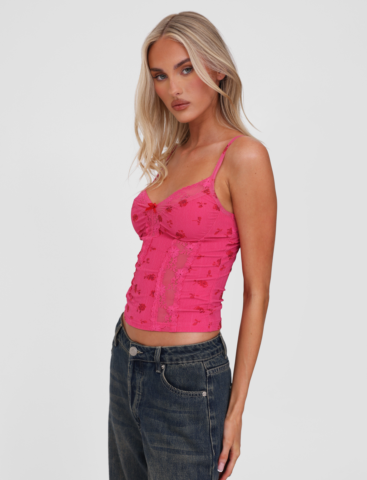 A woman with long blonde hair models the TIGERMIST KHALIA TOP - DITSY FLORAL, a fitted pink floral camisole with lace trim, paired with dark blue jeans and posed sideways against a plain white background.