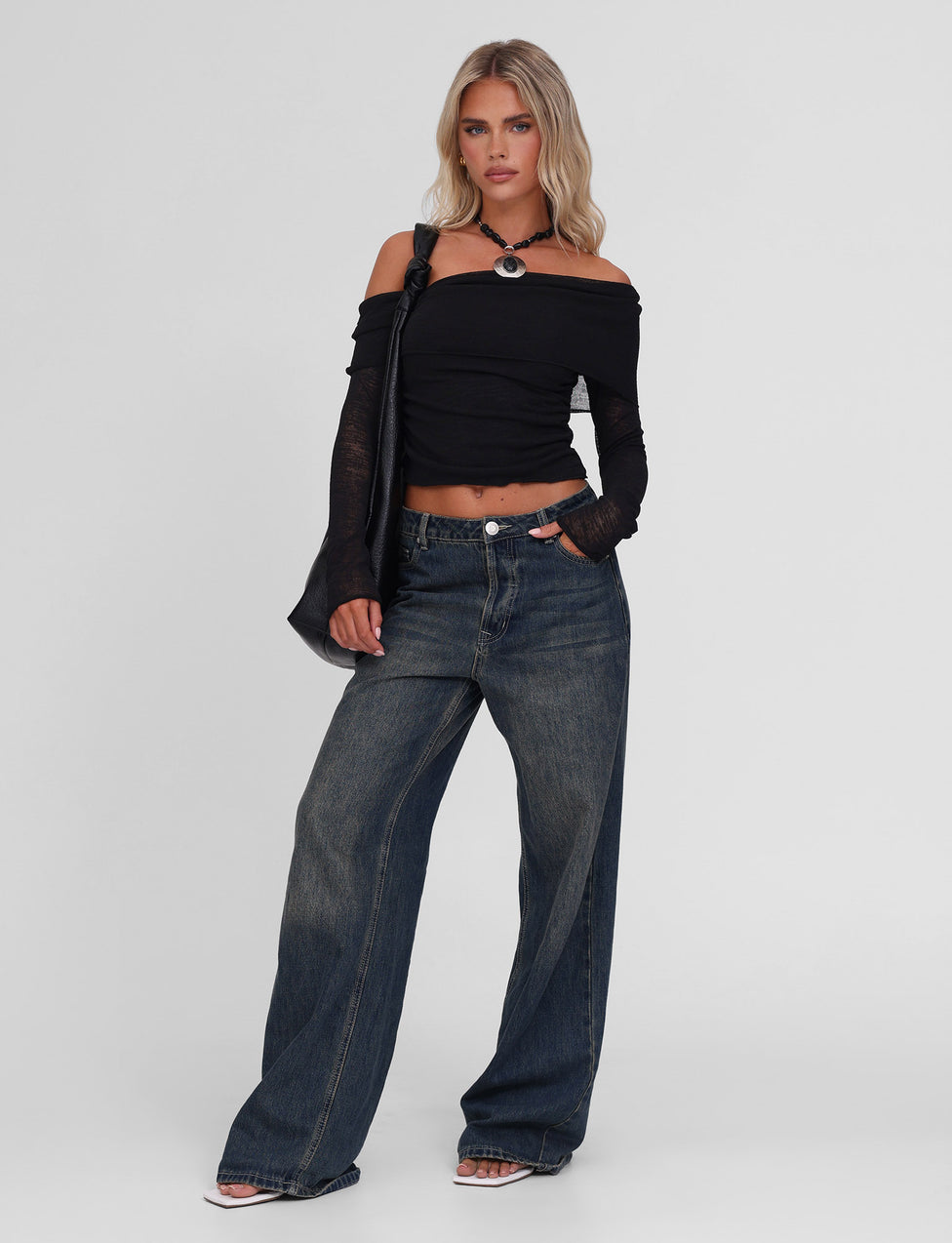 A blonde woman models the TIGERMIST VERA TOP - BLACK, a lightweight textured knit off-the-shoulder style, paired with wide-leg dark jeans and sandals. She carries a black shoulder bag and poses against a plain white background.