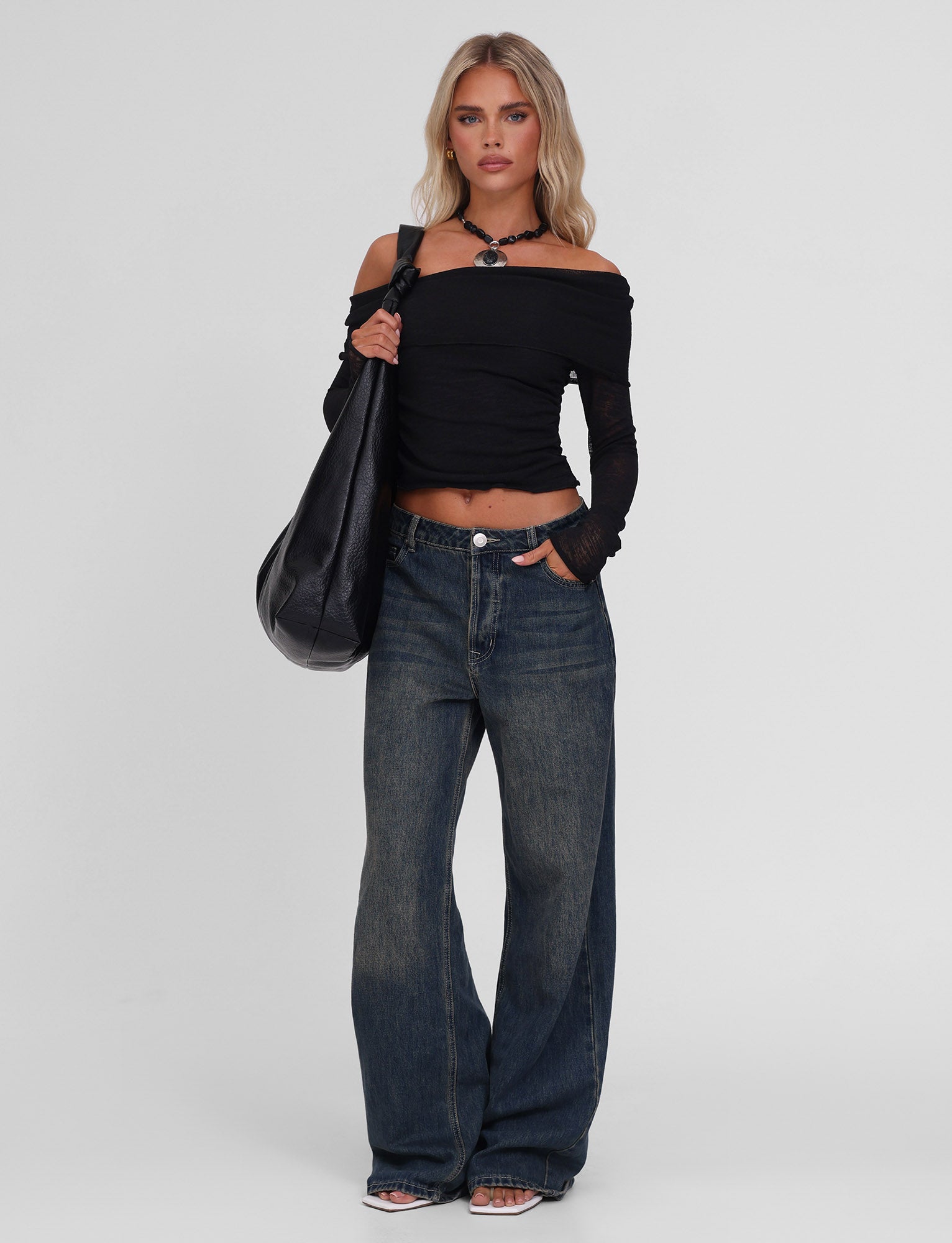A woman stands against a plain background wearing the TIGERMIST VERA TOP in black, loose blue jeans, black sandals, a black necklace, and carrying a large black bag over her shoulder.