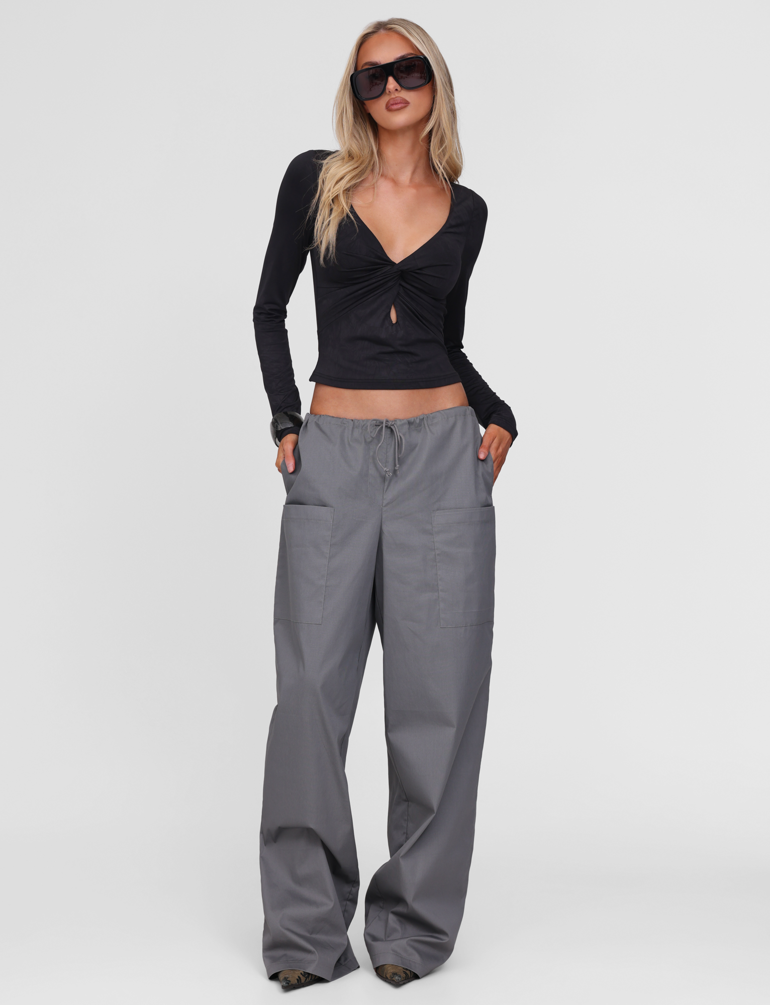 A woman stands with her hands in her pockets against a plain white background, wearing sunglasses, a long-sleeve black crop top with a front cut-out, and the TIGERMIST TYSHA PANT - CHARCOAL wide leg cargo pants with a drawstring waist.