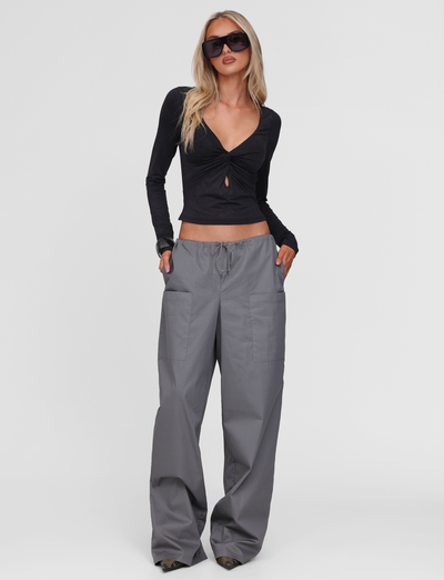 A woman stands with her hands in her pockets against a plain white background, wearing sunglasses, a long-sleeve black crop top with a front cut-out, and the TIGERMIST TYSHA PANT - CHARCOAL wide leg cargo pants with a drawstring waist.