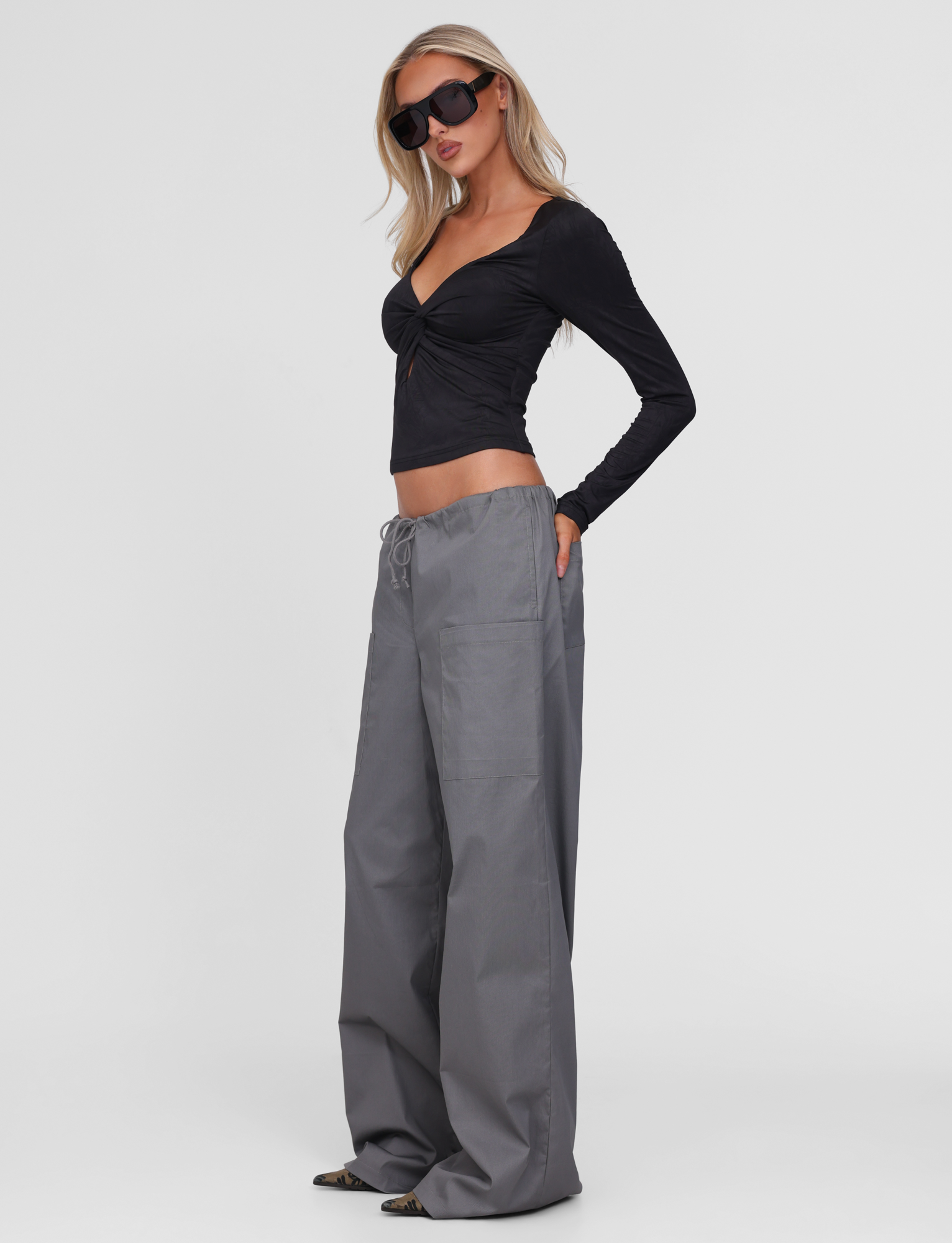 A woman stands confidently against a plain white background wearing dark sunglasses, a long-sleeve black crop top, and TIGERMIST's TYSHA PANT in charcoal with a drawstring waist and wide legs.