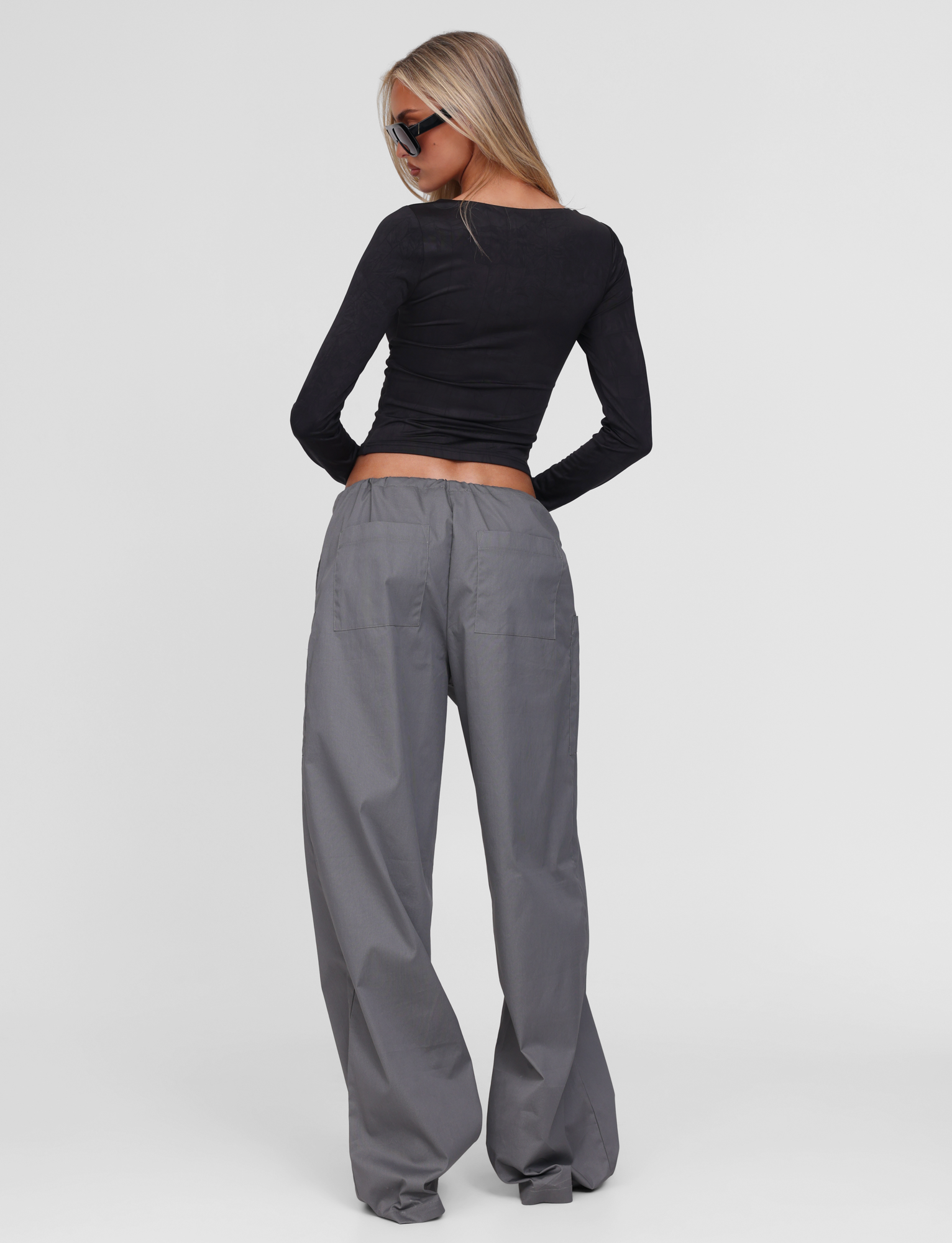 A woman with long blonde hair and black sunglasses wears the TIGERMIST TYSHA PANT in charcoal, featuring a drawstring waist, along with a fitted black long-sleeve top, standing with her back to the camera against a white background.