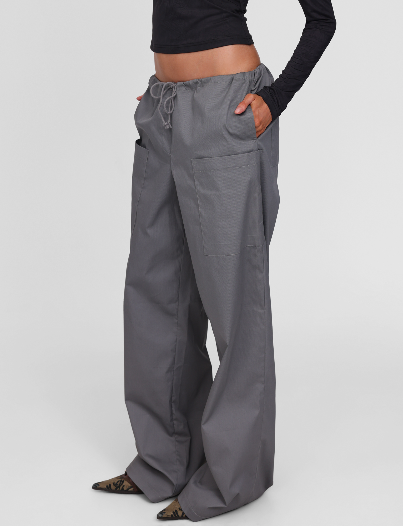 A person models the TIGERMIST TYSHA PANT in charcoal—wide leg cargo style with a drawstring waist and large front pockets—paired with a black long-sleeve top and pointed shoes, standing against a white background.
