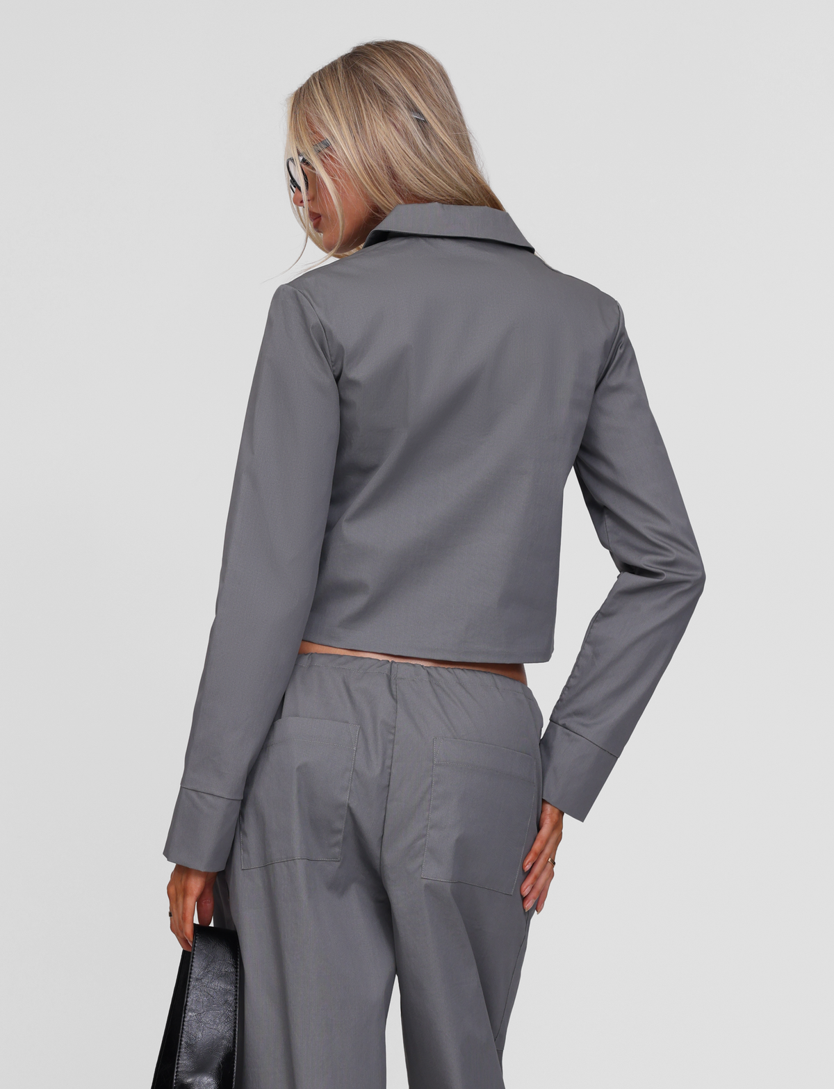 A woman with long blonde hair and sunglasses wears the TIGERMIST TYSHA JACKET in charcoal with matching pants, facing away from the camera and holding a black handbag in her left hand.