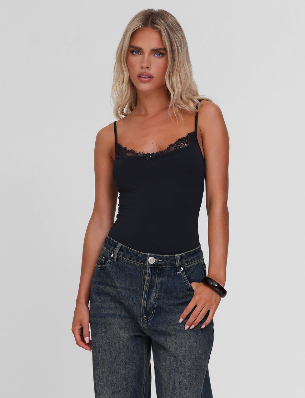 A woman with long blonde hair wears the TIGERMIST ASTRID BODYSUIT in black, featuring a lace trim and adjustable straps, paired with loose blue jeans. She stands against a plain light gray background with a neutral expression.