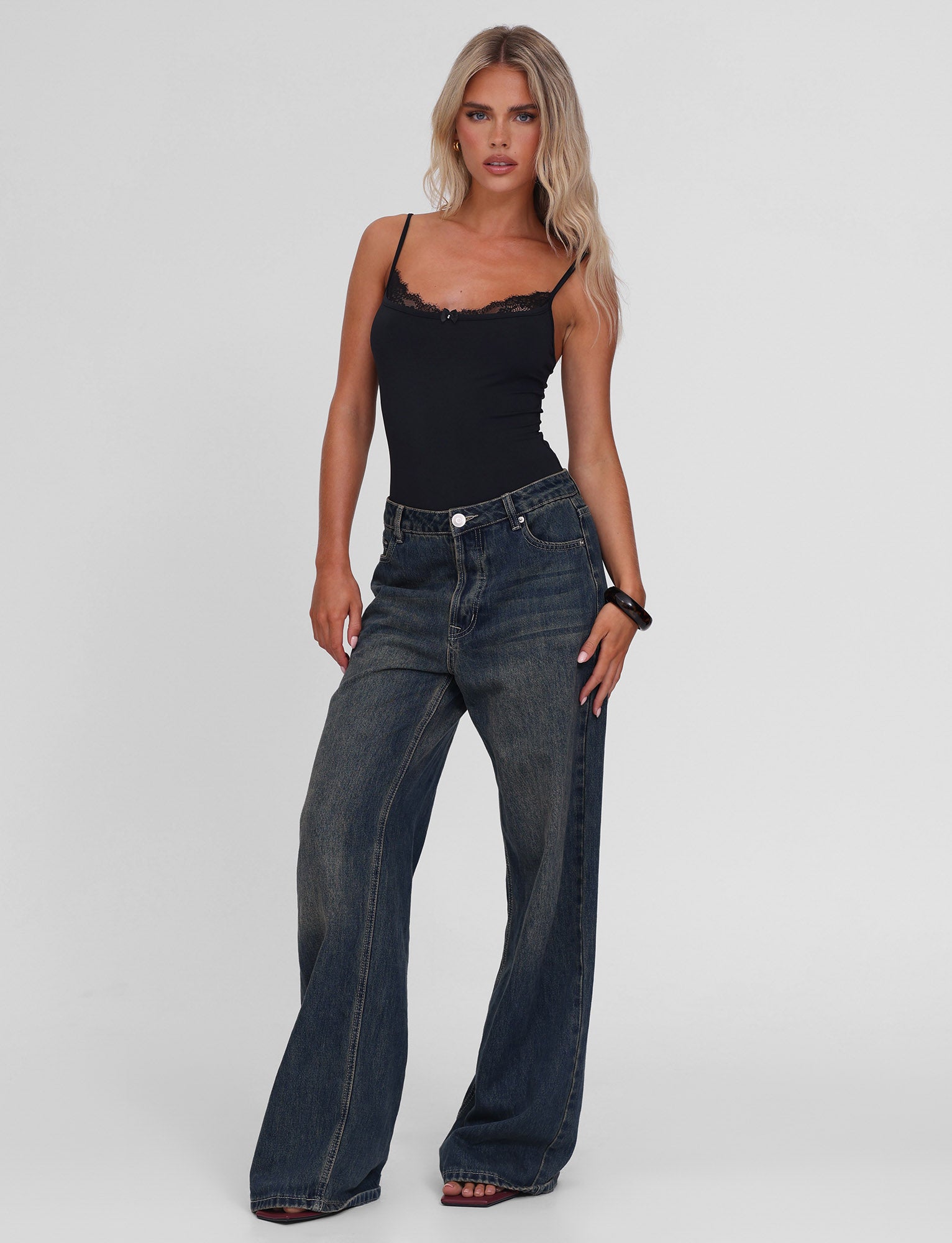 A woman with long blonde hair wears the TIGERMIST ASTRID BODYSUIT in black, featuring adjustable straps, paired with loose high-waisted dark blue jeans. She stands confidently against a plain white background.