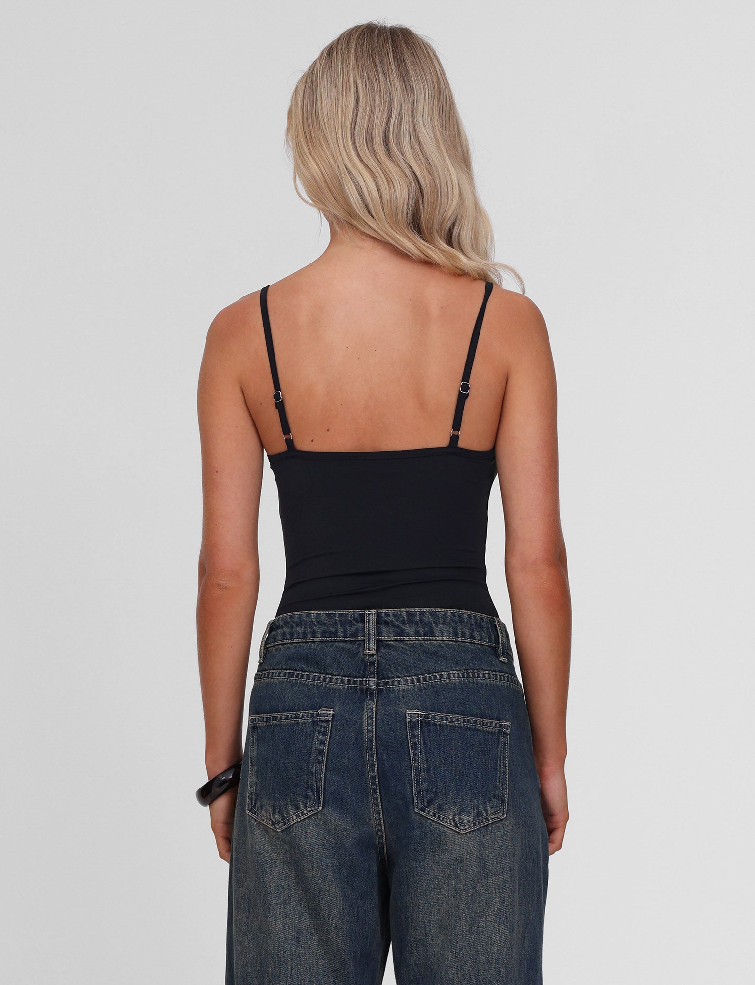 A person with long, wavy blonde hair is seen from the back wearing the TIGERMIST ASTRID BODYSUIT in black, featuring adjustable straps, paired with high-waisted blue jeans against a plain light gray background.