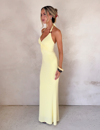 A woman in profile stands against a light textured wall, wearing the TIGERMIST ZENEVA MAXI DRESS in lemon. Her blonde hair is up, and she completes the elegant look with bracelets and earrings.