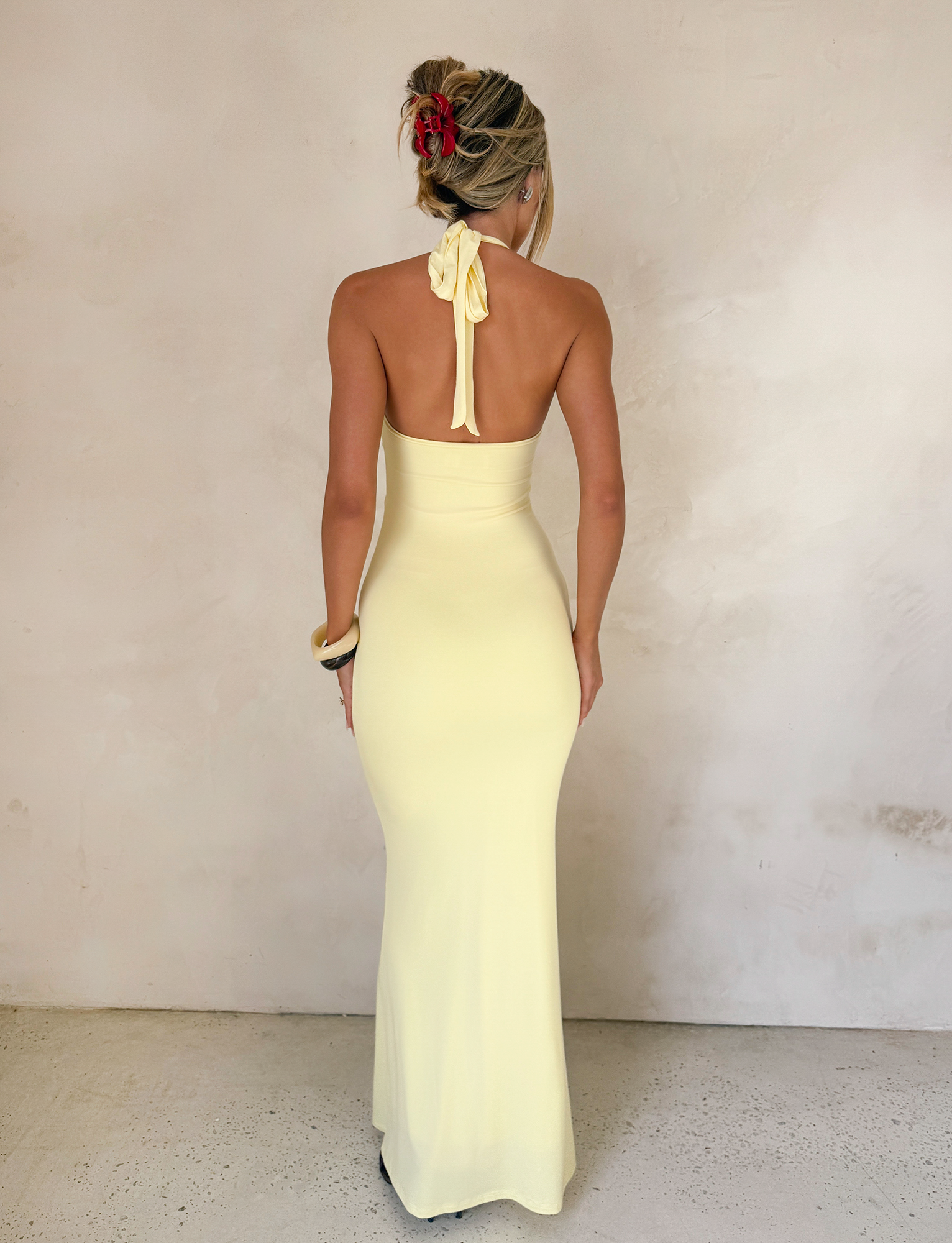 A blonde woman with a red hair accessory wears the TIGERMIST ZENEVA MAXI DRESS in Lemon, featuring a halter neckline and twist detail, photographed from behind against a textured off-white wall.