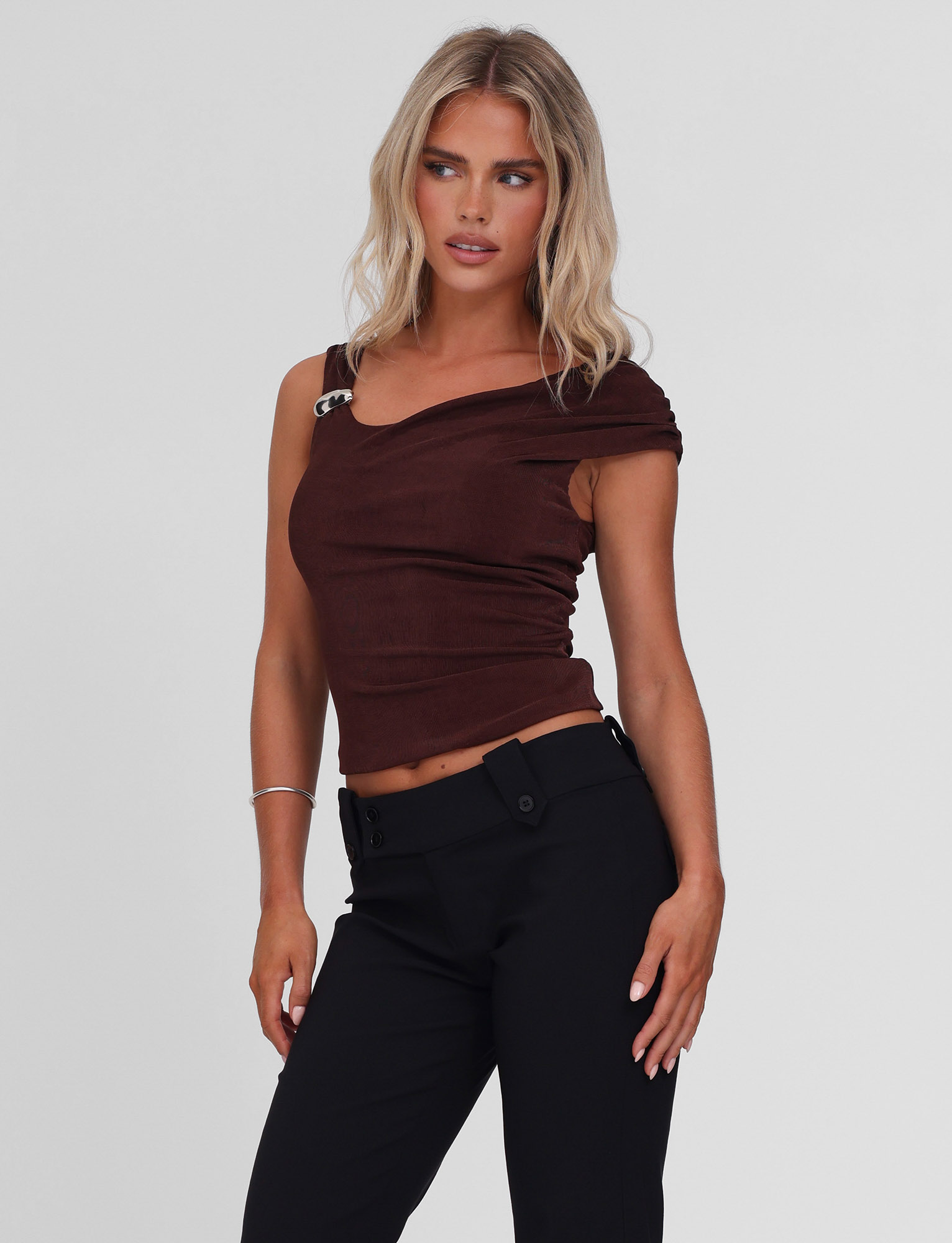 A woman with wavy blonde hair wears the TIGERMIST CASSIAN TOP in chocolate with an off-shoulder detail and silver clasp, paired with high-waisted black pants, standing against a plain light background, looking slightly to the side.