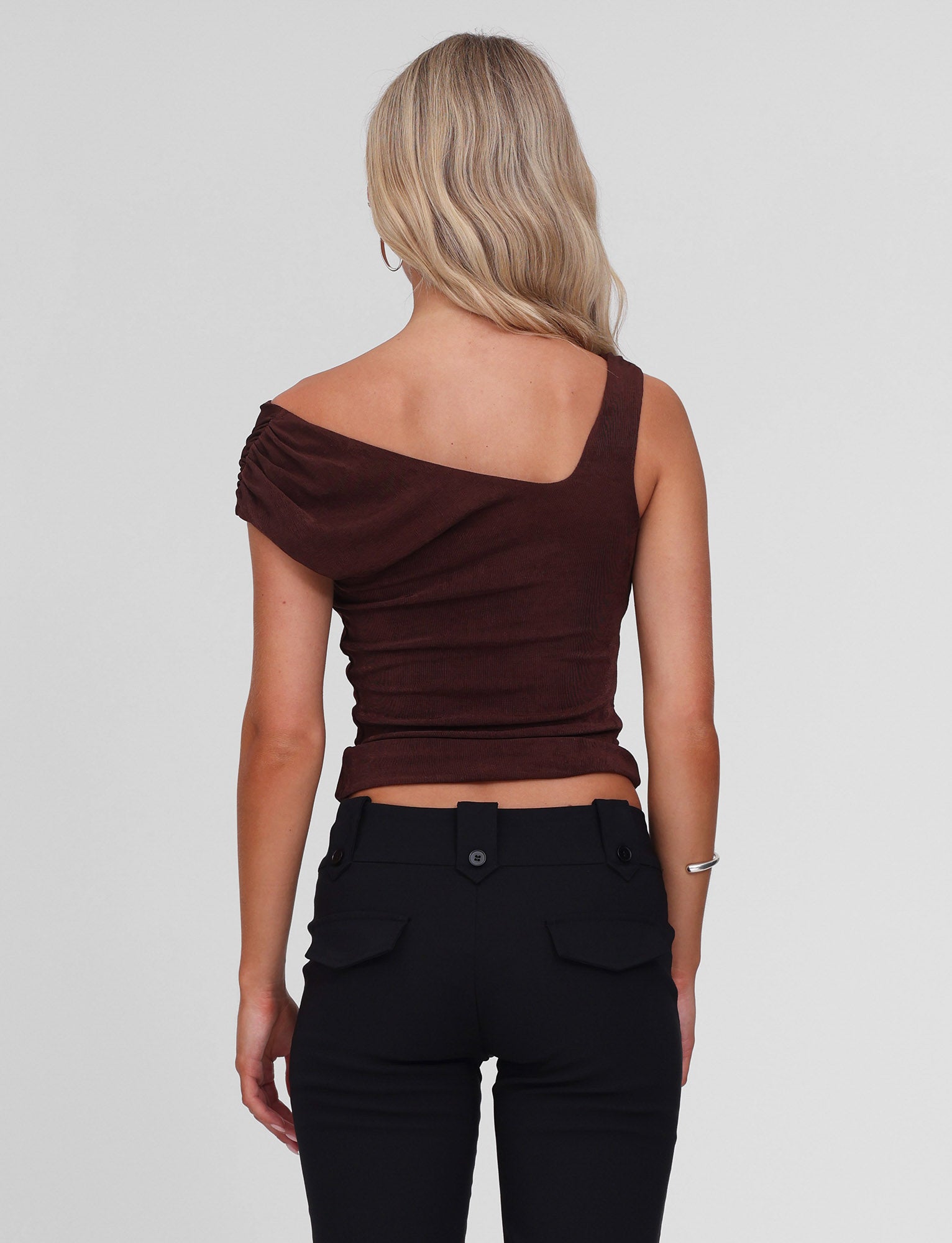 A woman with blonde wavy hair faces away, wearing the TIGERMIST CASSIAN TOP in chocolate and high-waisted black pants against a plain light background.