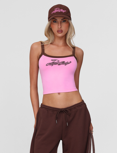 A woman in a pink tank top with brown straps and text, brown sweatpants, and the TIGERMIST MERRYN CAP in Chocolate stands with one hand in her pocket against a plain light gray background.