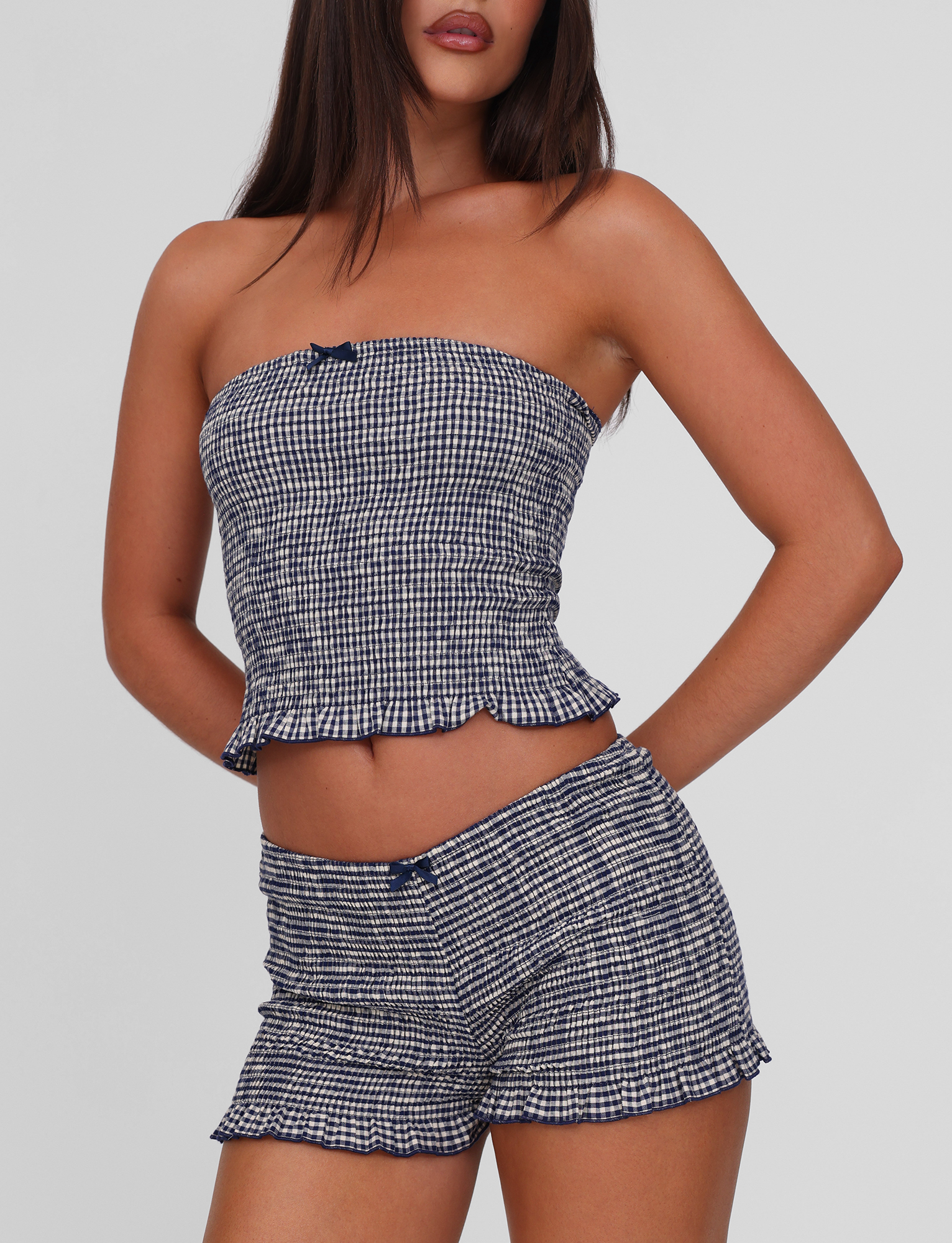 A woman poses with one hand on her hip, wearing TIGERMIST's ERYNN SHORTS in navy gingham and a matching strapless blue and white checkered top with a small center bow, set against a plain background.