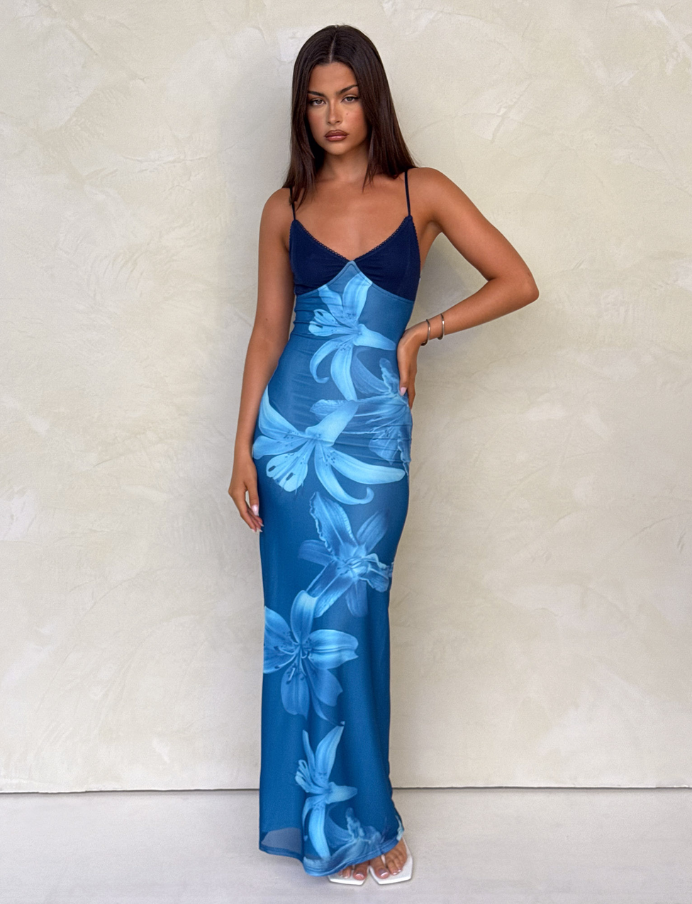 A woman wears the TIGERMIST ALAINA DRESS - BLUE ORCHID, a fully lined blue maxi with adjustable spaghetti straps and large floral prints, styled with white sandals as she stands against a textured wall looking at the camera.