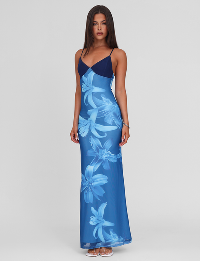 A woman with long straight hair and sandals poses against a plain background in the TIGERMIST ALAINA DRESS - BLUE ORCHID, a fully lined maxi dress featuring large light blue orchid floral prints and adjustable spaghetti straps.