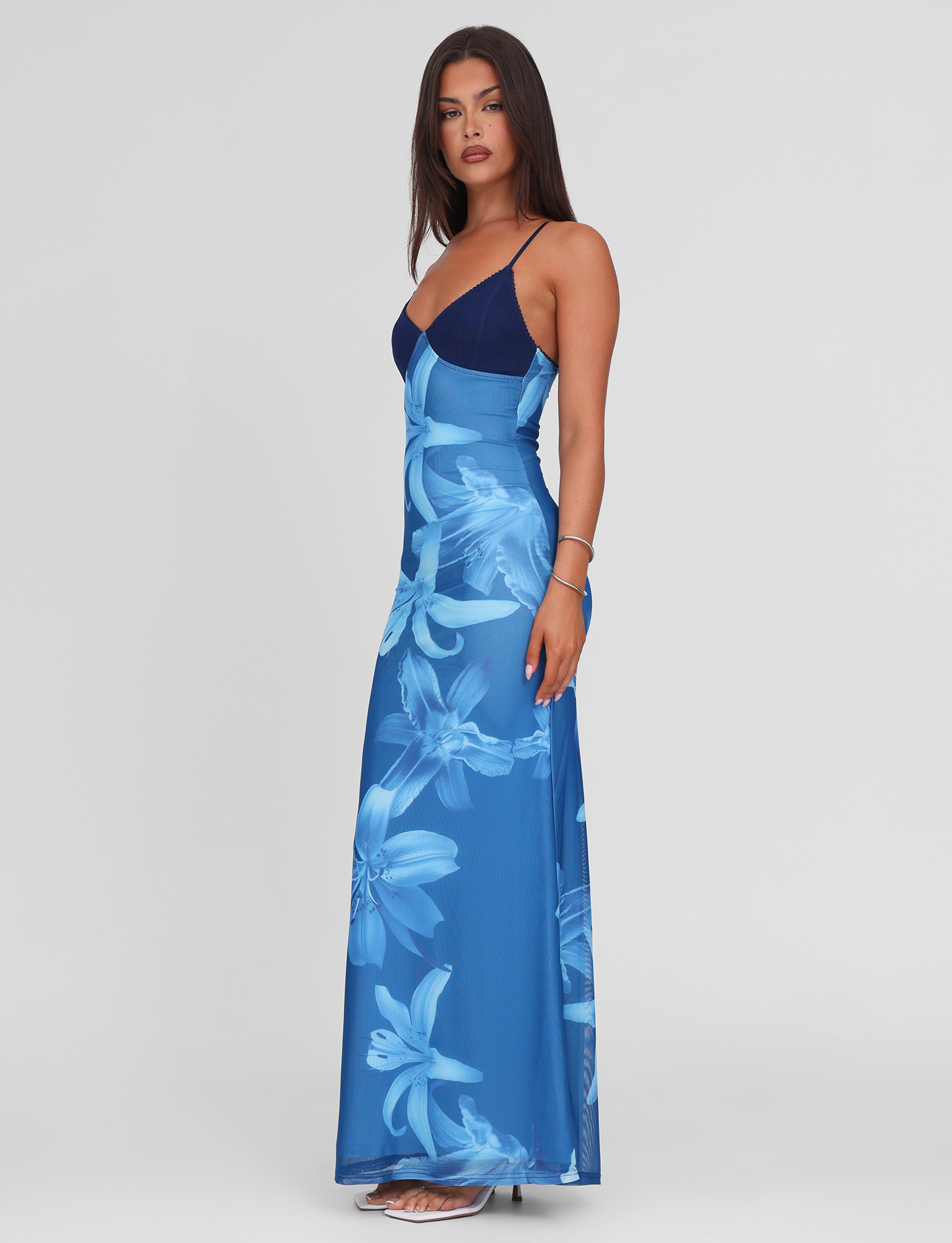 A woman with long dark hair poses sideways in the TIGERMIST ALAINA DRESS - BLUE ORCHID, a blue floral maxi with adjustable spaghetti straps and fitted bodice, against a plain white background. She is wearing sandals.