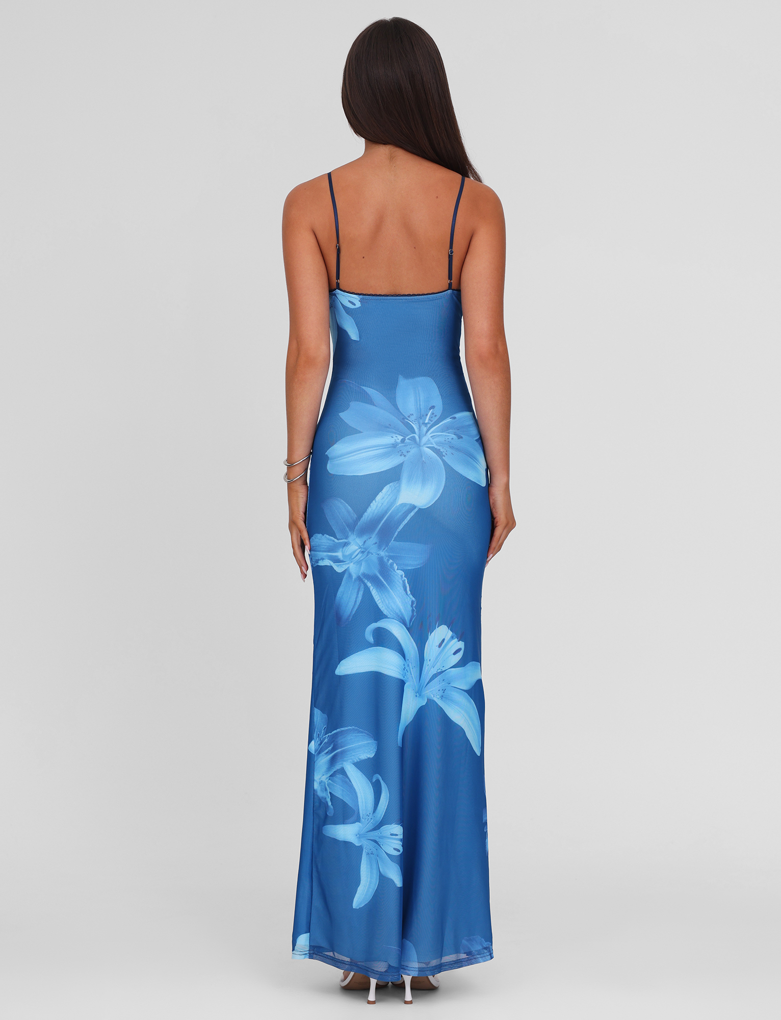 A woman with long dark hair, seen from behind, wears the TIGERMIST ALAINA DRESS - BLUE ORCHID, a blue maxi with adjustable spaghetti straps and large light blue blooms, standing against a plain light gray background.