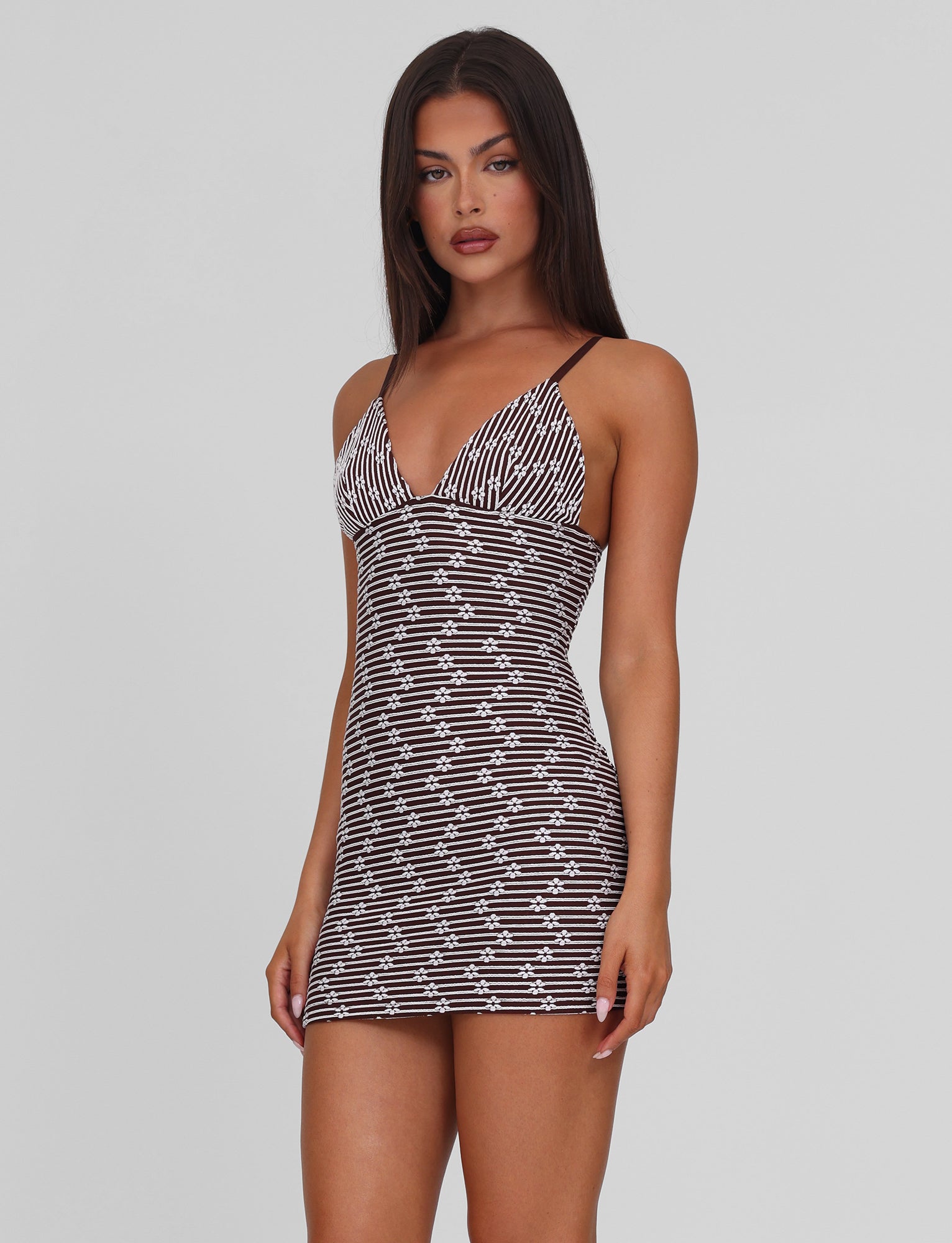 A woman with long dark hair wears the TIGERMIST SAHRA MINI DRESS in chocolate brown stripe, a fitted, sleeveless style with thin straps, standing against a plain light gray background.